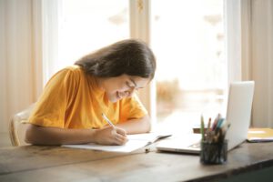 Studentin sitzt an Tisch und schreibt etwas auf. Vor ihr steht ein Laptop.